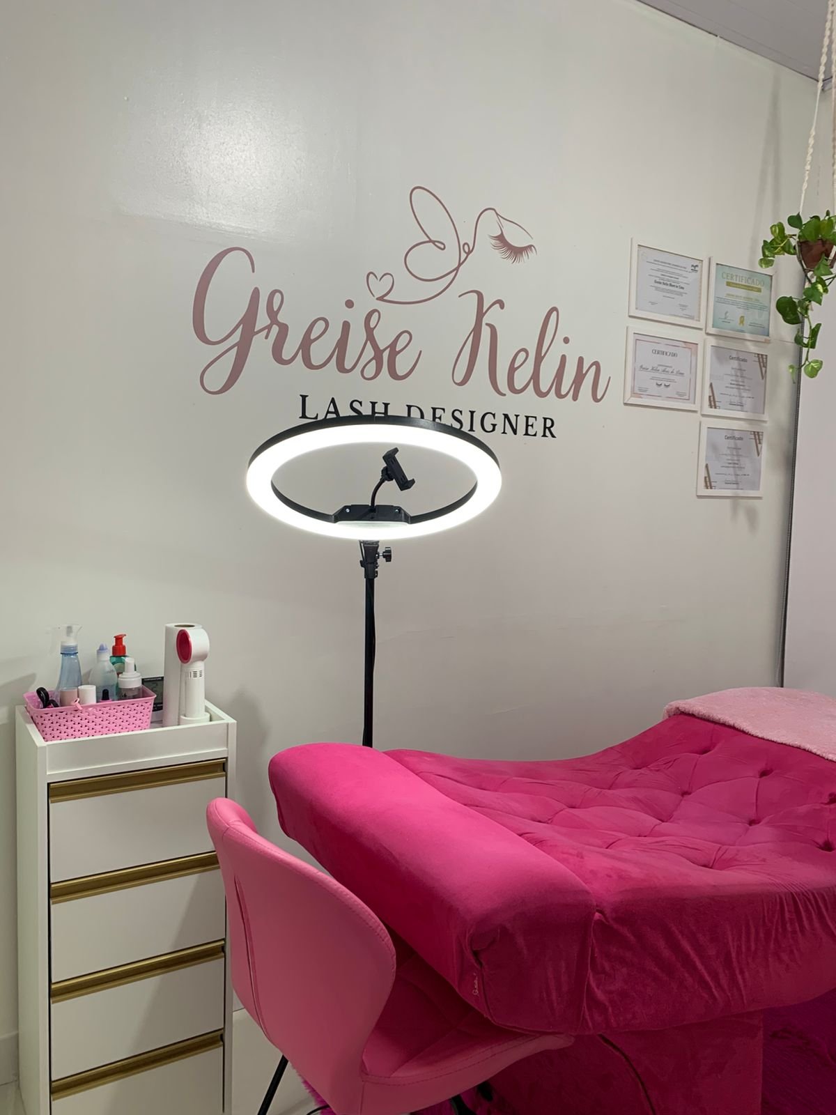 Detalhe do estúdio GK Lash & Beauty com maca rosa aveludada, cadeira rosa, iluminação de aro e prateleira com produtos ao lado dos certificados na parede