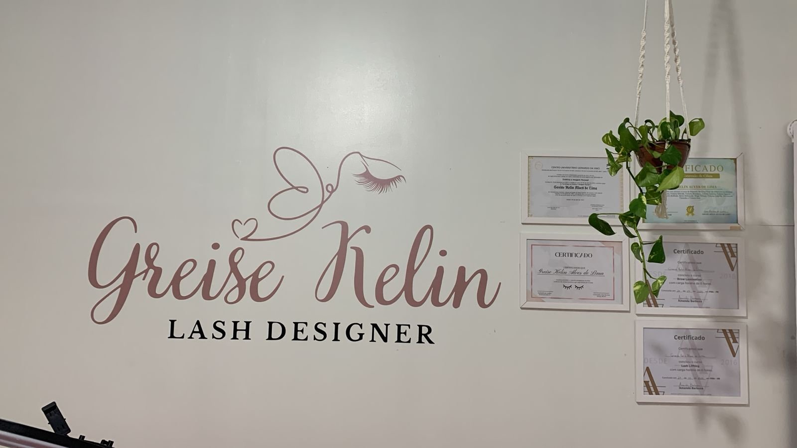 Parede do estúdio GK Lash & Beauty com logomarca Greise Kelin Lash Designer, certificados emoldurados e planta suspensa, evidenciando profissionalismo e ambiente acolhedor